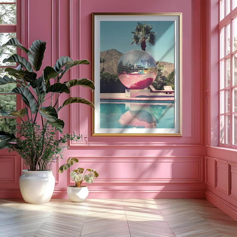 May include: A framed print of a large disco ball floating in a swimming pool. The disco ball is pink and silver and reflects the surrounding landscape. The print is set against a pink wall with white trim.