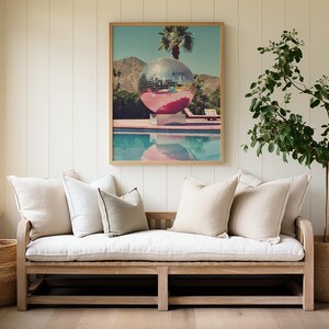 May include: A white couch with beige cushions sits in front of a white wall with wood paneling. A framed print of a disco ball in a pool hangs above the couch. A potted plant with green leaves sits in a woven basket to the right of the couch.
