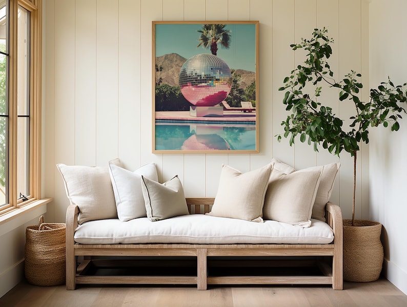 May include: A white couch with beige cushions sits in front of a white wall with wood paneling. A framed print of a disco ball in a pool hangs above the couch. A potted plant with green leaves sits in a woven basket to the right of the couch.