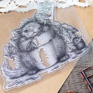 May include: A clear silicone rubber stamp featuring a detailed black ink illustration of a fluffy cat sitting inside a flower pot, surrounded by foliage, with a small sign above that reads CAT NIP and a tiny mouse standing to the right side.