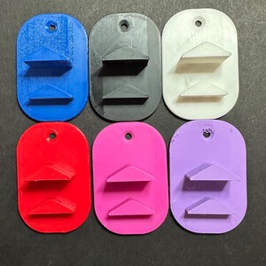 May include: Six oval-shaped wall mounts arranged in two rows of three. Each mount features two horizontal triangular ledges and a single circular hole at the top for mounting. The colors include blue, black, white, red, pink, and light purple.