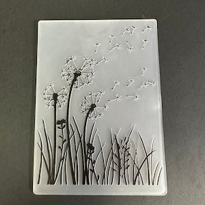 May include: A rectangular, translucent plastic craft stencil featuring a black dandelion design. The design includes three dandelion seed heads, grass, and seeds blowing in the wind. The stencil is designed for crafting and DIY projects.