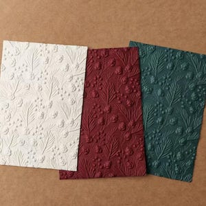 May include: Three rectangular sheets of paper in white, deep red, and dark green, each featuring an embossed pattern of pine needles, small berries, and pinecones covering the entire surface of the paper in a repeating botanical design.
