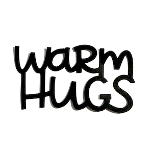 May include: Black wooden sign with the words "WARM HUGS" in a whimsical, handwritten font. The sign is set against a clean white background, creating a high-contrast visual. The letters have a slight shadow effect.