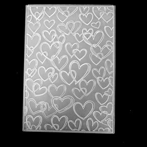 May include: A rectangular clear plastic embossing folder featuring a repeating pattern of etched heart outlines. The surface displays numerous overlapping heart shapes of varying sizes, creating a textured design across the entire transparent sheet.