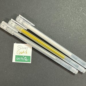 May include: Four gel pens with clear barrels and white caps. One pen has a gold ink, while the other three have silver ink. A small white card with the words "Silver, Gold, White" is also present.