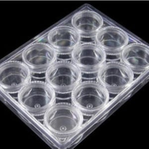 May include: A clear plastic storage container featuring twelve individual circular compartments arranged in a grid. Each compartment has a matching screw-top lid, providing a transparent view of the contents within this organized storage unit.
