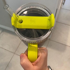 May include: A clear tumbler with a yellow lid and handle. The lid has a straw and the Stanley logo. The tumbler is held by a hand, and the background is a kitchen setting.