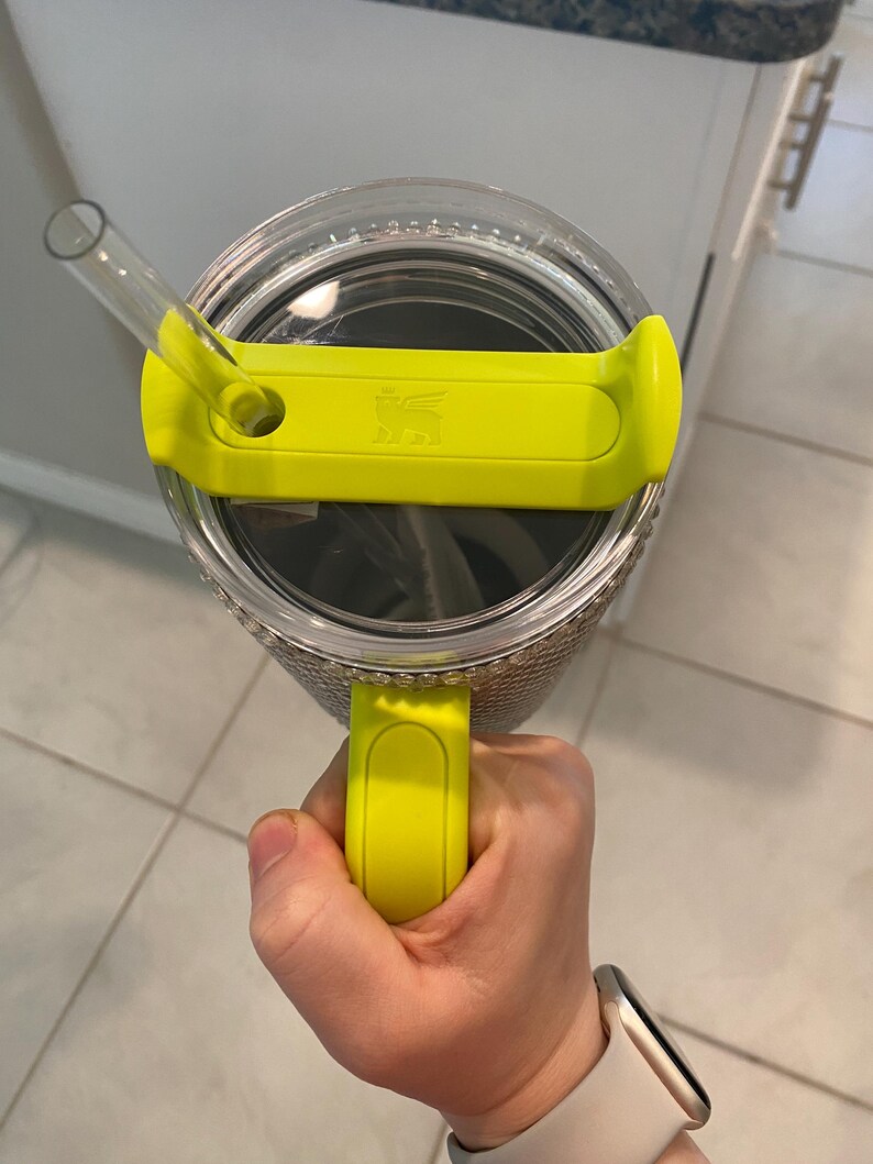 May include: A clear tumbler with a yellow lid and handle. The lid has a straw and the Stanley logo. The tumbler is held by a hand, and the background is a kitchen setting.