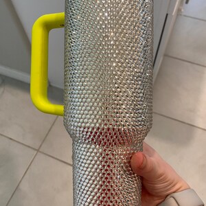 May include: A large, bedazzled tumbler with a silver rhinestone exterior. The tumbler has a bright yellow handle and a clear top. The bottom portion of the tumbler has a red interior. The tumbler is designed to hold a large quantity of liquid.