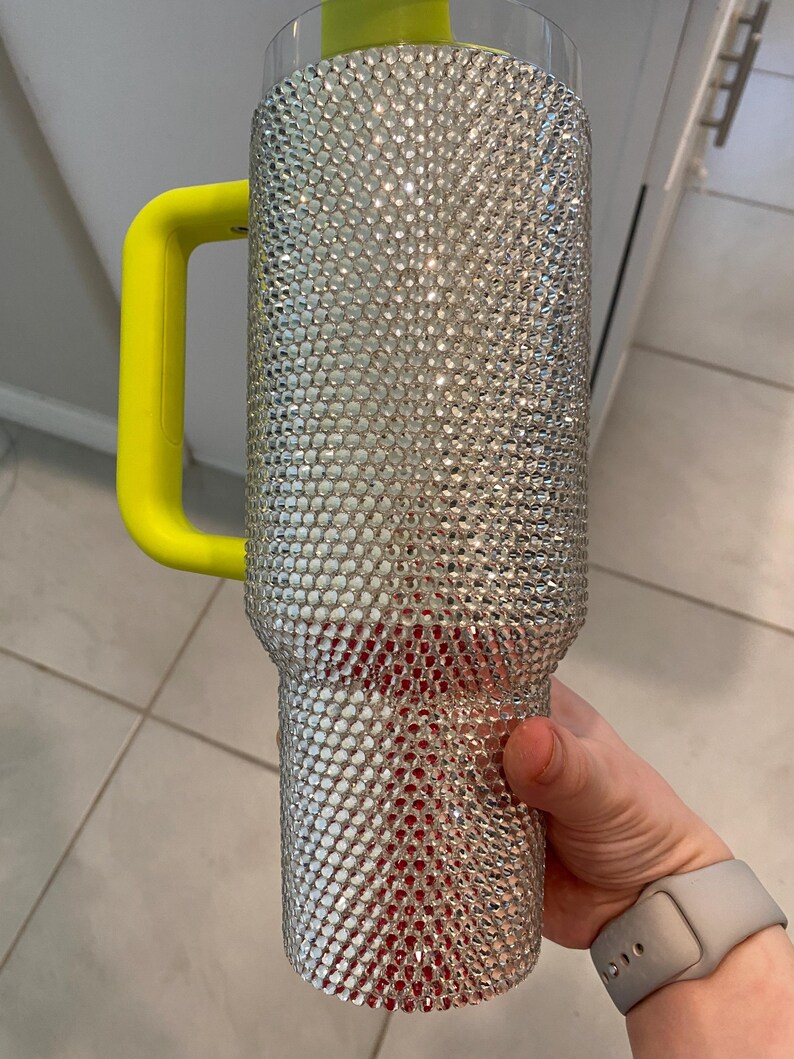 May include: A large, bedazzled tumbler with a silver rhinestone exterior. The tumbler has a bright yellow handle and a clear top. The bottom portion of the tumbler has a red interior. The tumbler is designed to hold a large quantity of liquid.