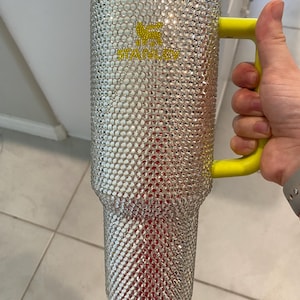 May include: A large, clear Stanley tumbler bedazzled with silver rhinestones. The tumbler has a yellow handle and lid. The Stanley logo is in yellow. The interior appears red. The tumbler is held by a person.