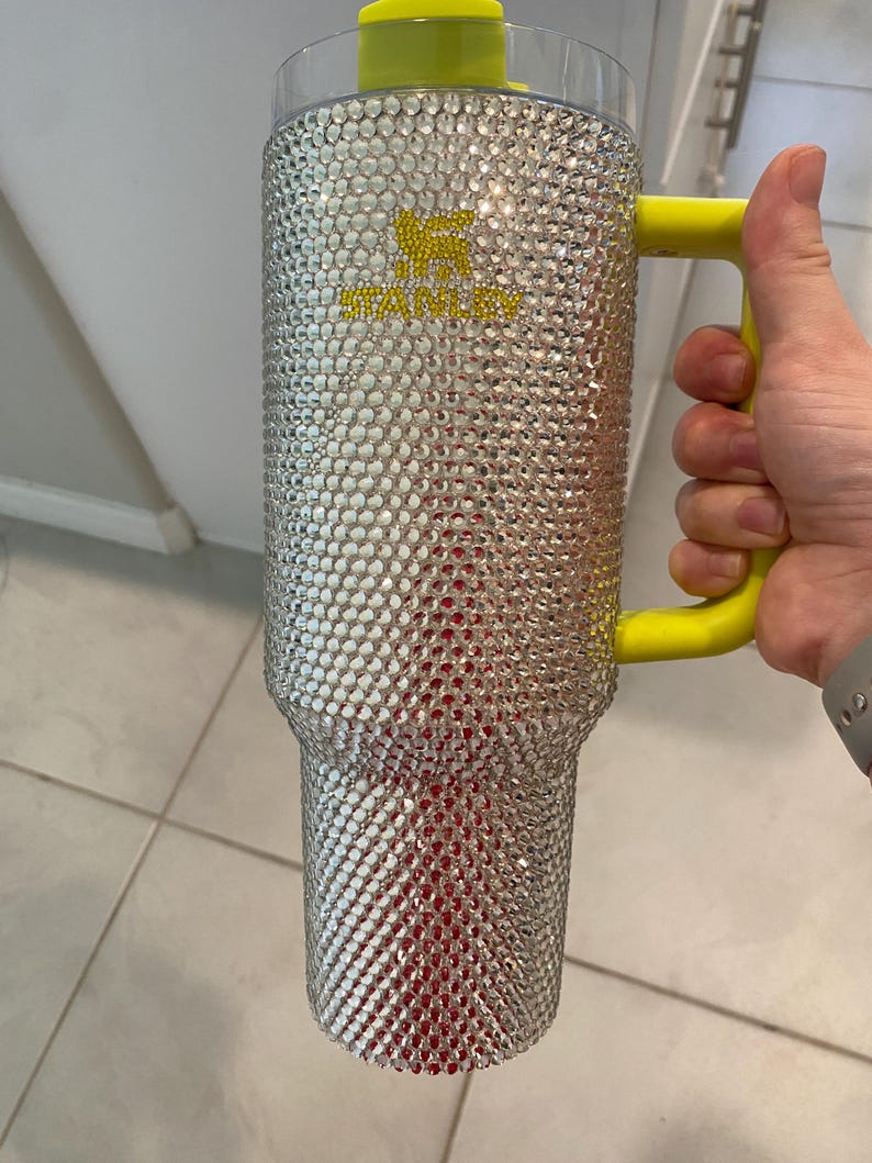 May include: A large, clear Stanley tumbler bedazzled with silver rhinestones. The tumbler has a yellow handle and lid. The Stanley logo is in yellow. The interior appears red. The tumbler is held by a person.