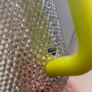 May include: A close-up of a silver-colored, bedazzled container with a bright yellow handle. The container is covered in small, clear, sparkling rhinestones. The handle is a solid, curved shape. The text "H.S.S. By Hale" is visible.
