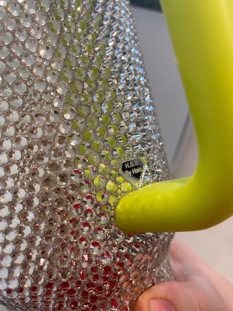 May include: A close-up of a silver-colored, bedazzled container with a bright yellow handle. The container is covered in small, clear, sparkling rhinestones. The handle is a solid, curved shape. The text "H.S.S. By Hale" is visible.