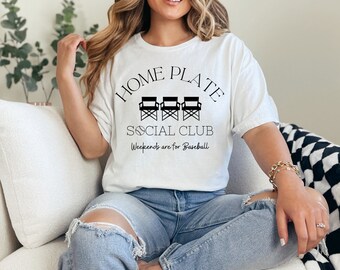 Op de afbeelding: Wit T-shirt met zwarte tekst die "Home Plate Social Club" en "Weekends are for Baseball" luidt. De tekst wordt omringd door een cirkel met drie zwarte regisseursstoelen erin.