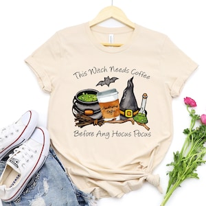 May include: Beige t-shirt with the text "This Witch Needs Coffee Before Any Hocus Pocus". The graphic includes a witch's hat, cauldron, coffee cup, and a bat. White sneakers and blue jeans are also visible.