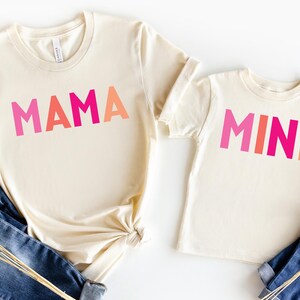 May include: Two cream colored t-shirts with the words "MAMA" and "MINI" printed in pink, orange, and red.