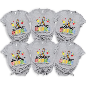 May include: Six gray t-shirts with a colorful puzzle piece ribbon and the text "Support Squad" in black. Each shirt has a different arrangement of colorful blocks with the letters "S-U-P-P-O-R-T" and "S-Q-U-A-D" printed on them. The hashtag "#TeamCustom" is printed below the text on each shirt.