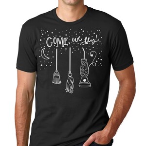 May include: A black short-sleeved t-shirt featuring a white graphic print of a crescent moon, stars, two brooms, and a vacuum cleaner. The text "COME, we fly!" is printed in a stylized white script font above the illustrated objects.