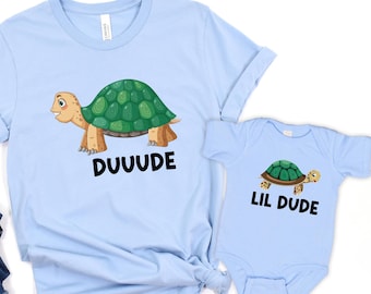 May include: Light blue t-shirts and a baby onesie featuring a cartoon turtle design. The adult shirt has the word "DUUUDE" printed below the turtle, while the onesie says "LIL DUDE".