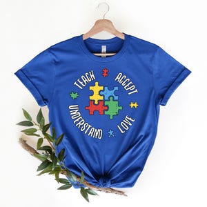 May include: Royal blue t-shirt with a white circle graphic featuring a puzzle piece design and the text "Teach Accept Understand Love".