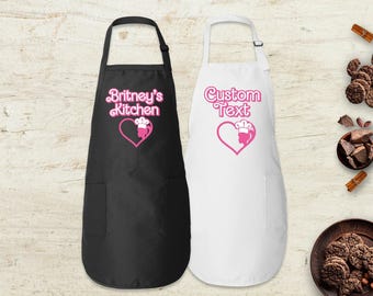 May include: A black apron with the white text Britney's Kitchen and a pink heart icon featuring a chef hat, alongside a white apron with the pink text Custom Text and a matching pink heart icon with a chef hat, both featuring two front pockets.