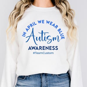 May include: White sweatshirt with blue text that reads "IN APRIL WE WEAR BLUE Autism AWARENESS #TeamCustom". The sweatshirt is long-sleeved and is paired with blue jeans.
