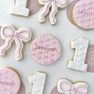 May include: A selection of decorated sugar cookies, including the number "1" and bow shapes, with pink and white icing. Some cookies are round and feature the name "Savannah". The cookies are arranged on a white surface.