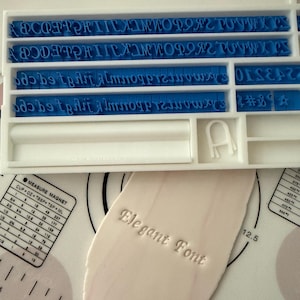 May include: A white plastic tray containing blue alphabet and number stamps. The stamps are arranged in rows and are used for creating impressions in clay or fondant. The tray also includes a ruler and a measurement chart.
