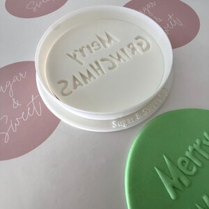 May include: A white plastic cookie cutter with the words "Merry Grinchmas" in a circular design. A green cookie with the same text is next to the cutter. The background includes pink oval stickers with the text "Sugar & Sweet".