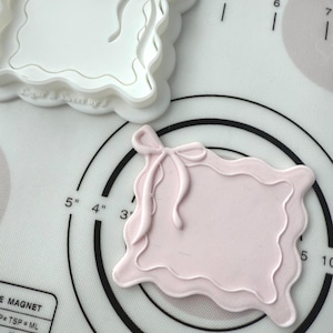 May include: A white and a pink cookie cutter with a bow design. The white cutter is next to a pink cookie with the same bow design. The text "Sugar & Sweet by" is visible on the white cutter. The image is on a white surface with black markings.