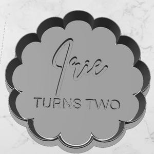 May include: A grey scalloped circular cookie cutter featuring the embossed text Irie in a cursive script font above the block capital text TURNS TWO. The tool has a raised outer edge and a flat interior surface for creating impressions in dough.