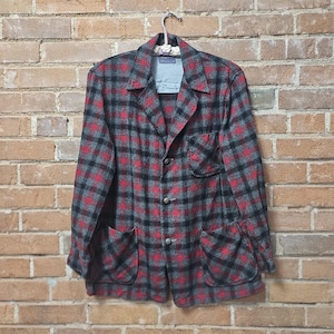 Può includere: Una giacca vintage a quadri rossi, neri e grigi è appesa a un muro di mattoni. La giacca presenta un classico design con bottoni, due tasche anteriori e un colletto a revers. Il motivo a quadri è audace e accattivante.