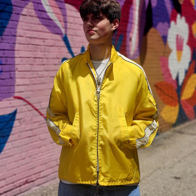 May include: A bright yellow jacket with a full zipper closure, white stripes on the sleeves, and two front pockets. The jacket is worn over a white t-shirt. The background features a colorful mural.
