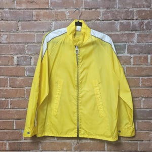 May include: A yellow zip-up jacket with white shoulder accents, hanging on a metal hanger. The jacket has a stand-up collar, two front pockets, and a full-length zipper. The jacket is made of a lightweight material.