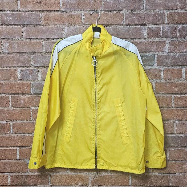 May include: A yellow zip-up jacket with white shoulder accents, hanging on a metal hanger. The jacket has a stand-up collar, two front pockets, and a full-length zipper. The jacket is made of a lightweight material.