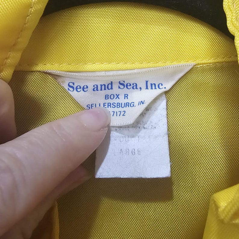 May include: Close-up of a yellow shirt collar with a white tag. The tag reads "See and Sea, Inc." with the address "BOX R SELLERSBURG, IN 47172" and the size "LARGE".