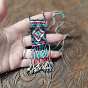 May include: A beaded necklace with a rectangular pendant featuring a diamond pattern in red, white, and turquoise on a navy blue background. The pendant has beaded fringe in turquoise, red, and silver. The necklace is held in a hand.