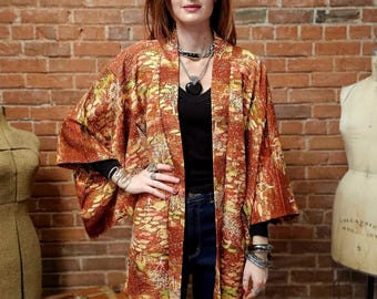 May include: A long, open-front kimono in shades of rust, yellow, and green. The kimono features a patterned design and wide sleeves. The garment is worn over a black top and blue jeans. The background is a brick wall.