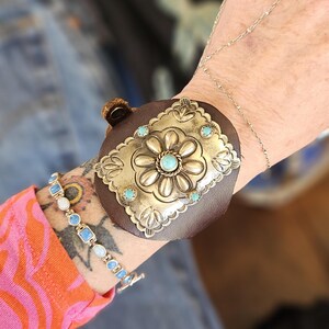 May include: A brown leather cuff bracelet with a silver-colored floral design and turquoise accents. The cuff features a rectangular silver plate with a flower in the center and smaller turquoise stones around the edges. A silver bracelet is also visible.