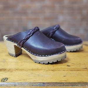 May include: A pair of dark purple clogs with a braided strap across the top. The clogs have a wooden sole and a thick, light-colored wooden heel. The soles have a textured, rubber-like bottom. White stitching accents the edges of the leather upper.