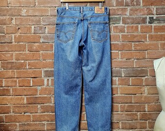 May include: Light blue denim jeans hanging on a wooden hanger against a brick wall. The jeans have a classic five-pocket design, with a small red tag on the back pocket. The jeans are a medium wash and appear to be vintage.