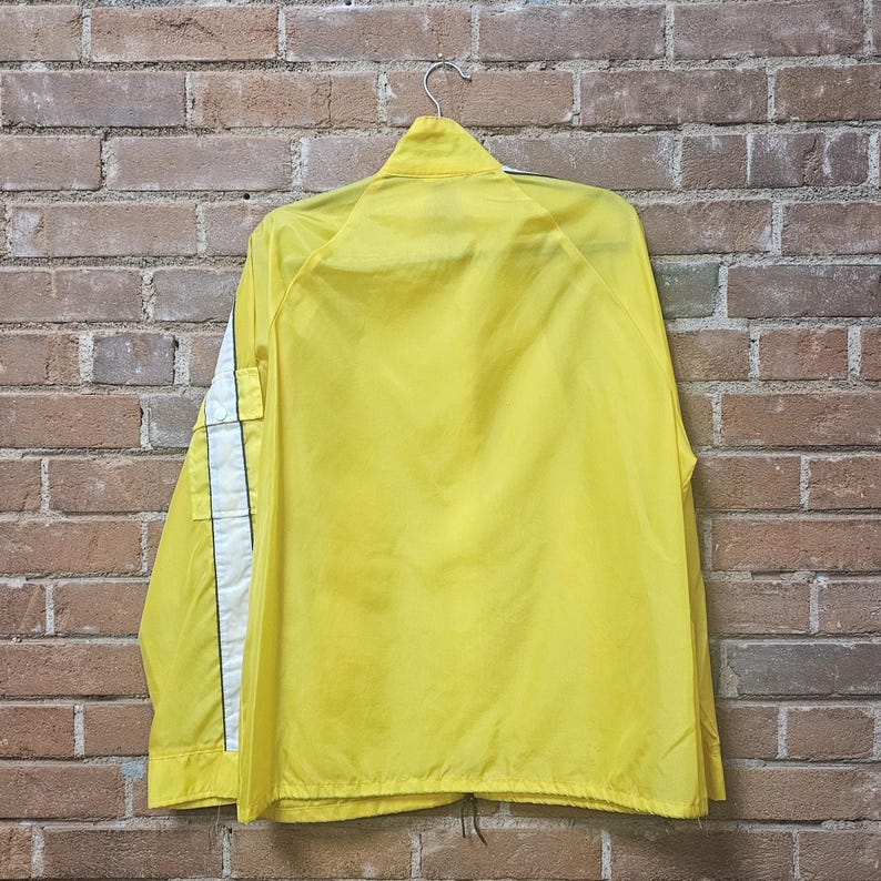 May include: A bright yellow jacket with a zipper closure and a collar, hanging against a brick wall. The jacket has white stripes down the sleeves and a black trim. The jacket is made of a lightweight material.