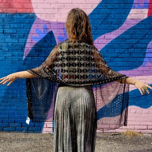 May include: A black sheer cape with a geometric silver pattern is draped over a silver pleated skirt. The cape has a delicate, open weave design. The background is a colorful mural with blue, pink, and purple paint.