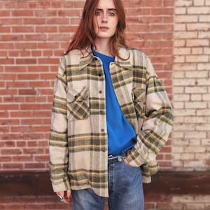 Può includere: Una camicia a quadri beige, verde e gialla con bottoni e due tasche frontali. La camicia è indossata sopra una t-shirt blu girocollo e jeans blu. Lo sfondo è un muro di mattoni rossi.