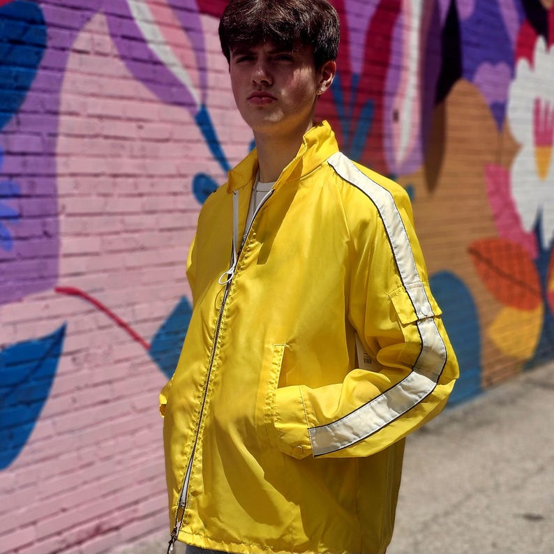 May include: A bright yellow jacket with white stripes down the sleeves and along the shoulders. The jacket has a zipper closure and two pockets. The background features a colorful mural.