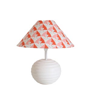 Può includere: Una lampada da tavolo bianca con paralume fantasia. Il paralume presenta un motivo ripetuto di uccelli bianchi su sfondo rosa e corallo. La base della lampada è una sfera bianca con una superficie strutturata e scanalata.