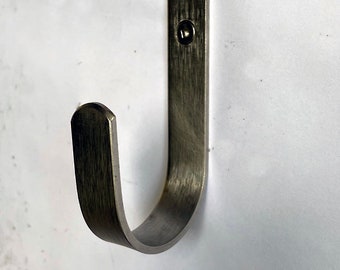 Puede incluir: Un gancho de metal plateado con la parte superior redondeada y un diseño en forma de J. El gancho está fijado a una pared con dos tornillos.