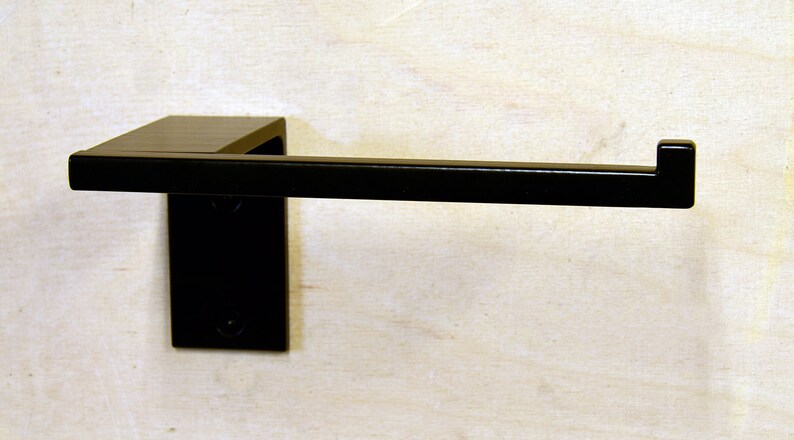 May include: A black metal toilet paper holder mounted to a wall. The holder has a square base and a rectangular arm.